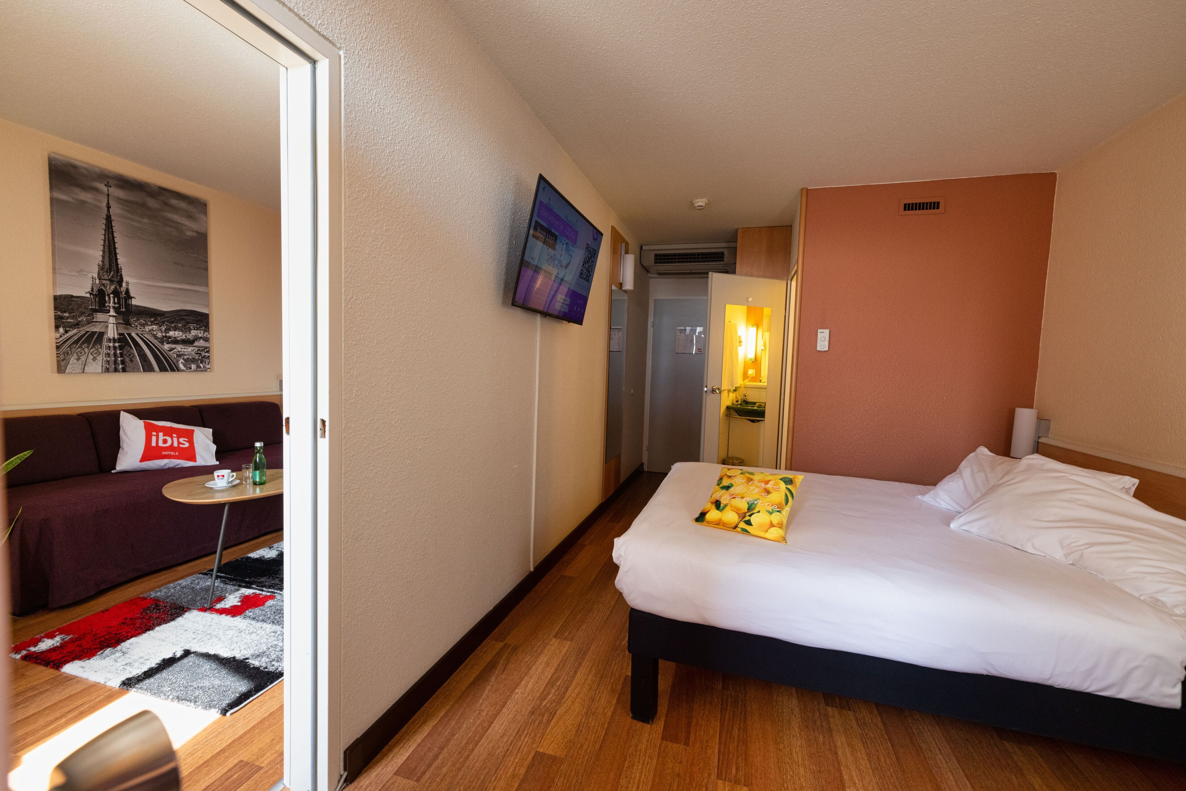 Een hotelkamer met een bed, een bank, een kleintje tafeltje met een kopje en een fles, een tv aan de muur en een schilderij van een gebouw.