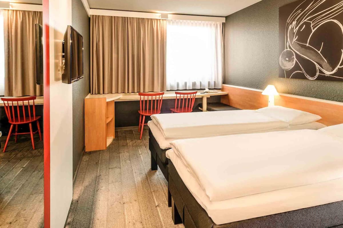 A hotel room with two beds, a desk, two red chairs, a TV, and a window with curtains.
