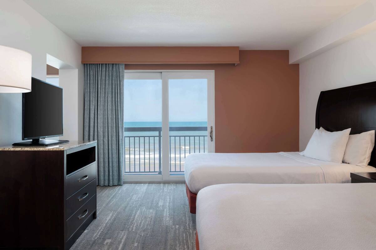 A hotel room with two beds, a TV, a desk, and a view of the ocean.