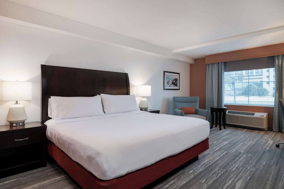Habitación de hotel con una cama grande, dos mesitas de noche con lámparas, una silla, una pequeña mesa y una ventana con cortinas.