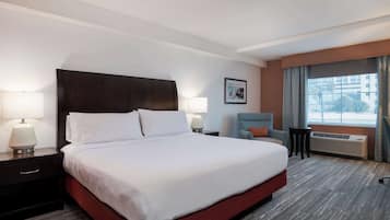 Habitación de hotel con una cama grande, dos mesitas de noche con lámparas, una silla, una pequeña mesa y una ventana con cortinas.