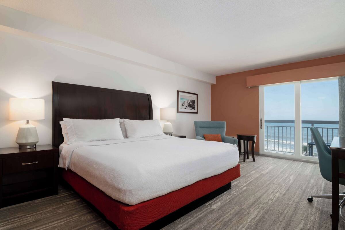 Habitación de hotel con una cama grande, dos mesitas de noche con lámparas, una silla, una pequeña mesa y un amplio ventanal con vista a la playa.