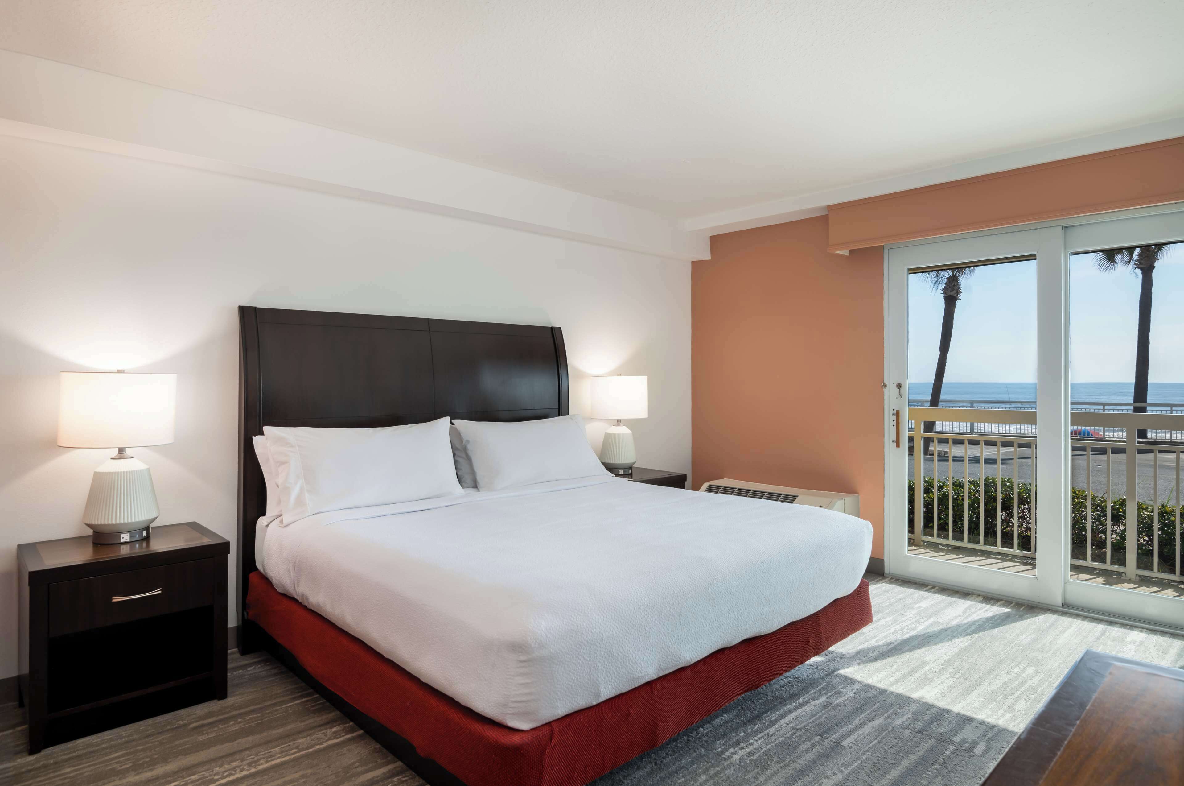 Een hotelkamer met een groot bed, twee nachtkastjes met lampen, uitzicht op de oceaan door een glazen schuifdeur en een balkon met een hekwerk.