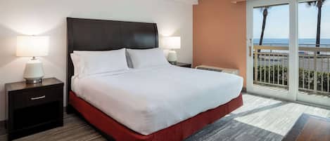 Een hotelkamer met een groot bed, twee nachtkastjes met lampen, uitzicht op de oceaan door een glazen schuifdeur en een balkon met een hekwerk.