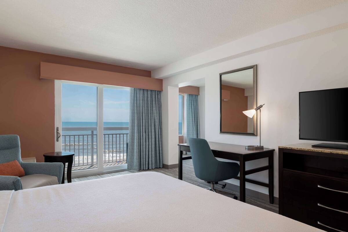 Habitación de hotel con cama, escritorio, silla, televisor y balcón con vista al océano.