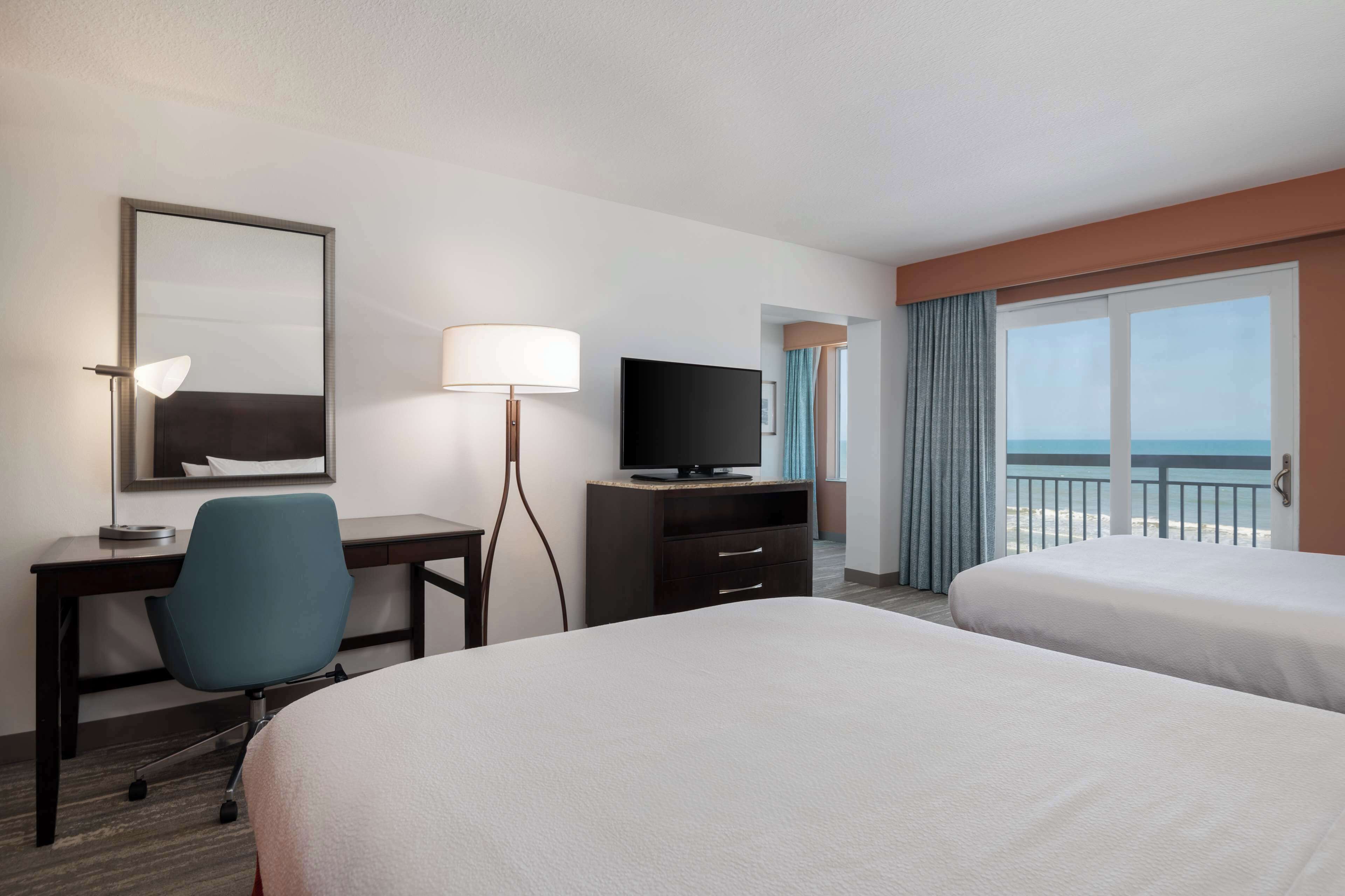 A hotel room with two beds, a desk with a chair, a television, and a large window with a view of the sea.