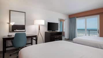 A hotel room with two beds, a desk with a chair, a television, and a large window with a view of the sea.