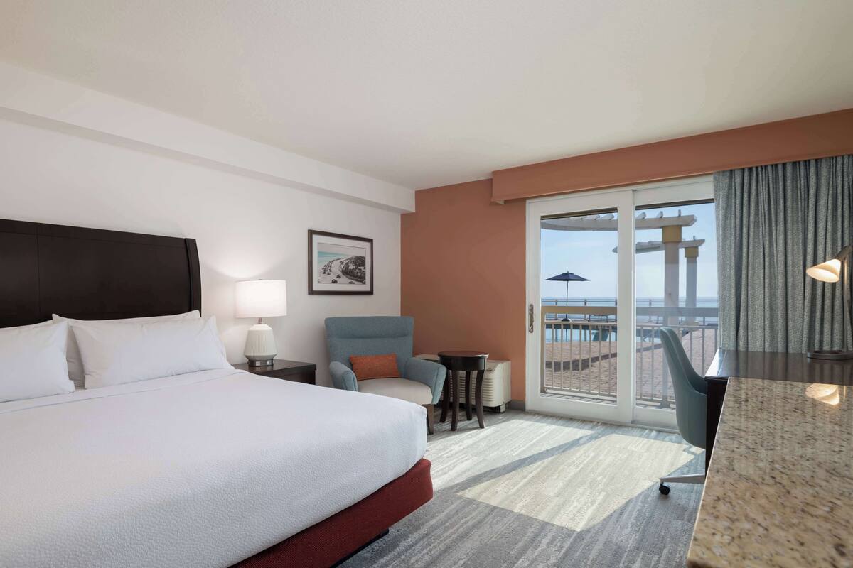 A hotel room with a large bed, a desk, a chair, a small table, and a balcony with a view.
