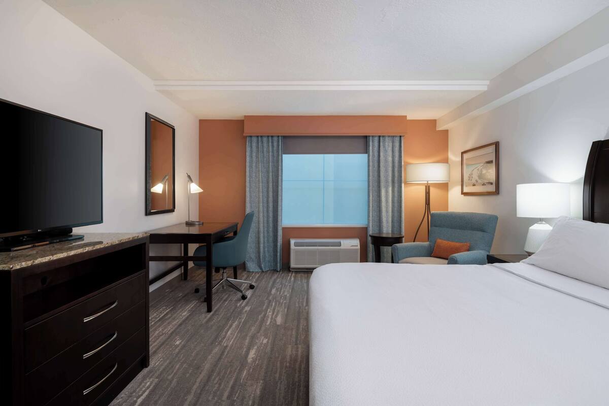 Habitación de hotel con cama, escritorio, silla, televisor y un amplio ventanal con cortinas.