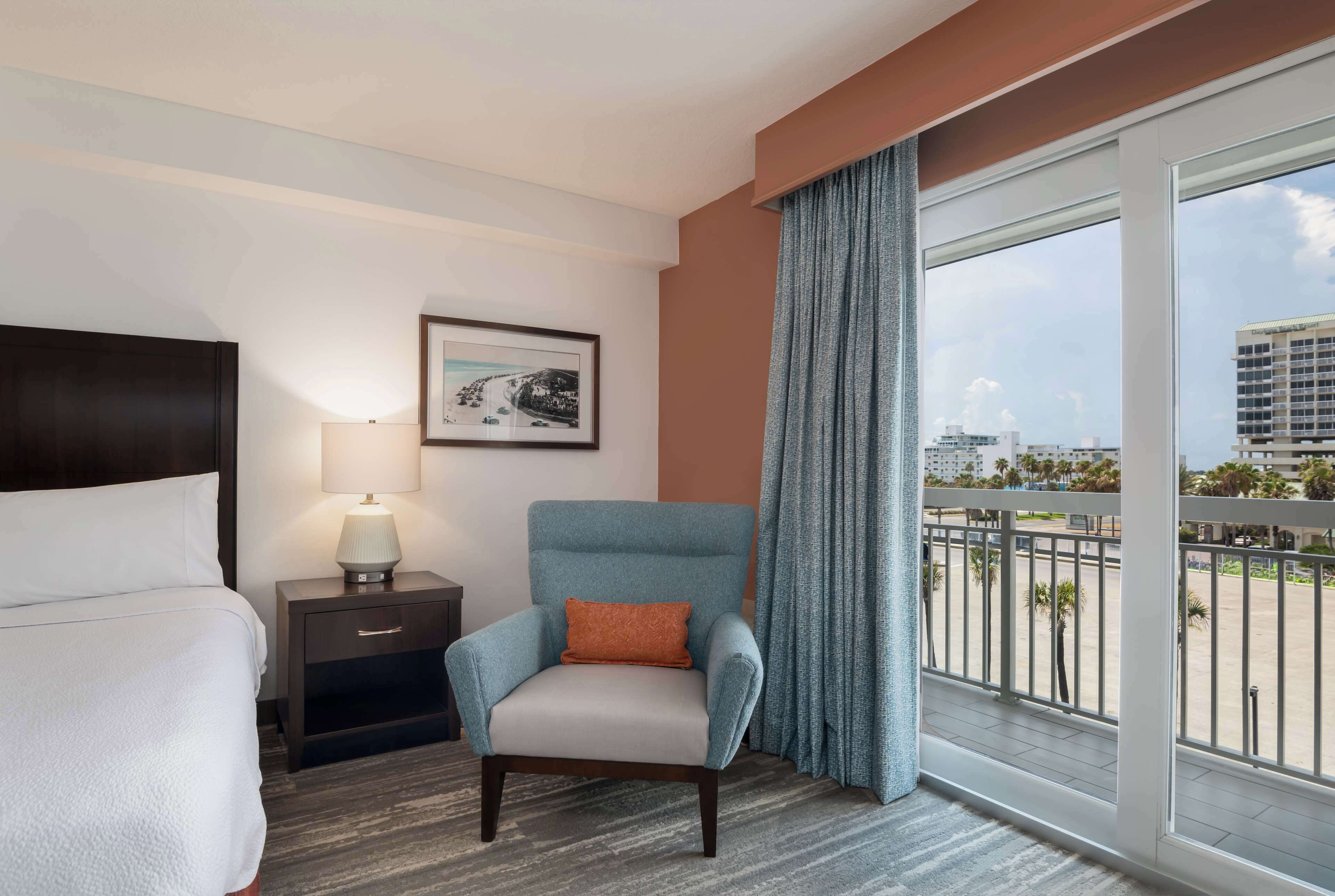 Habitación de hotel con cama, una silla, una mesita de noche con lámpara, un cuadro en la pared y un ventanal con vistas a edificios y palmeras.