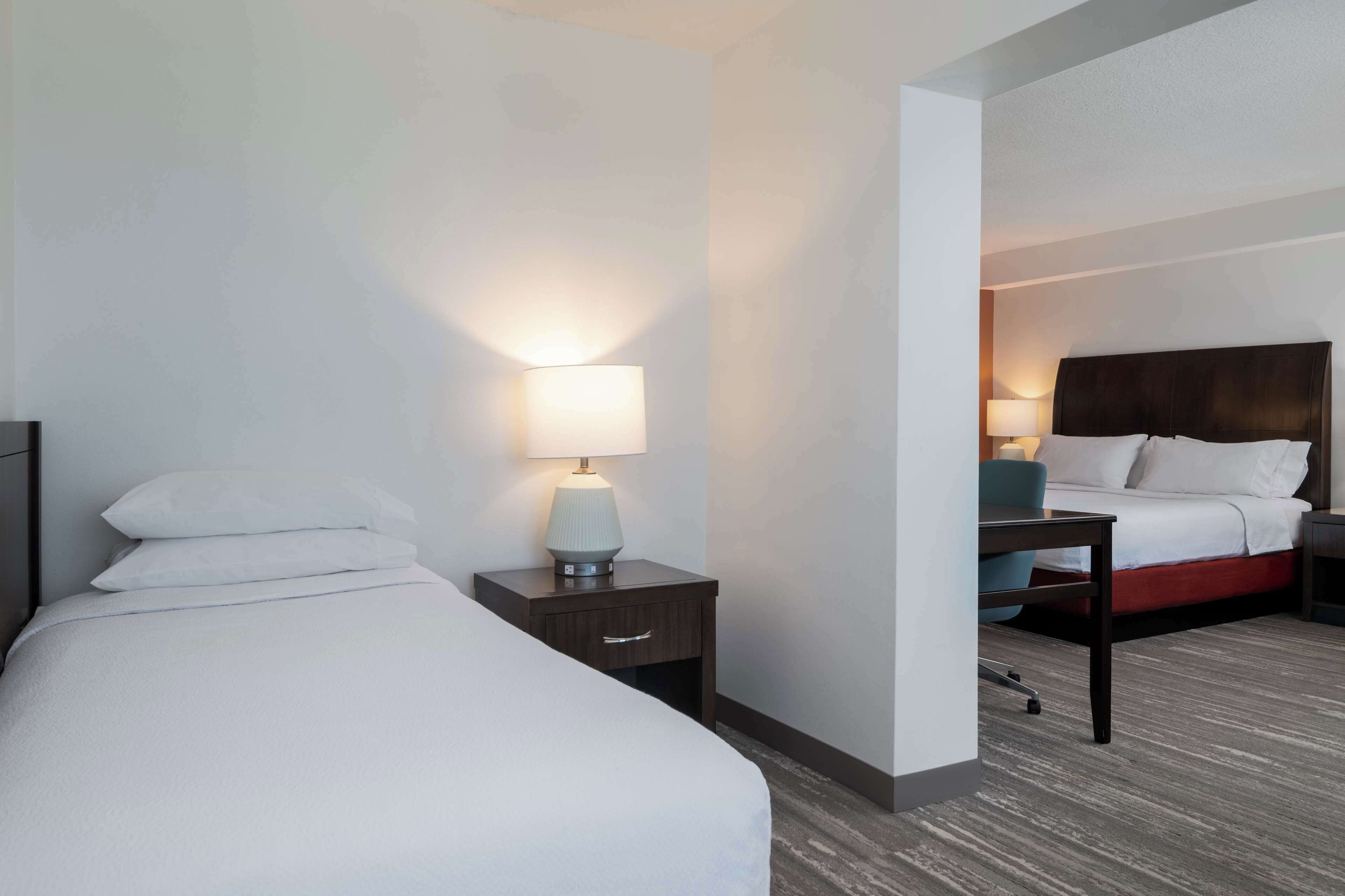 A hotel room with a bed, a nightstand with a lamp, a desk, and a chair.