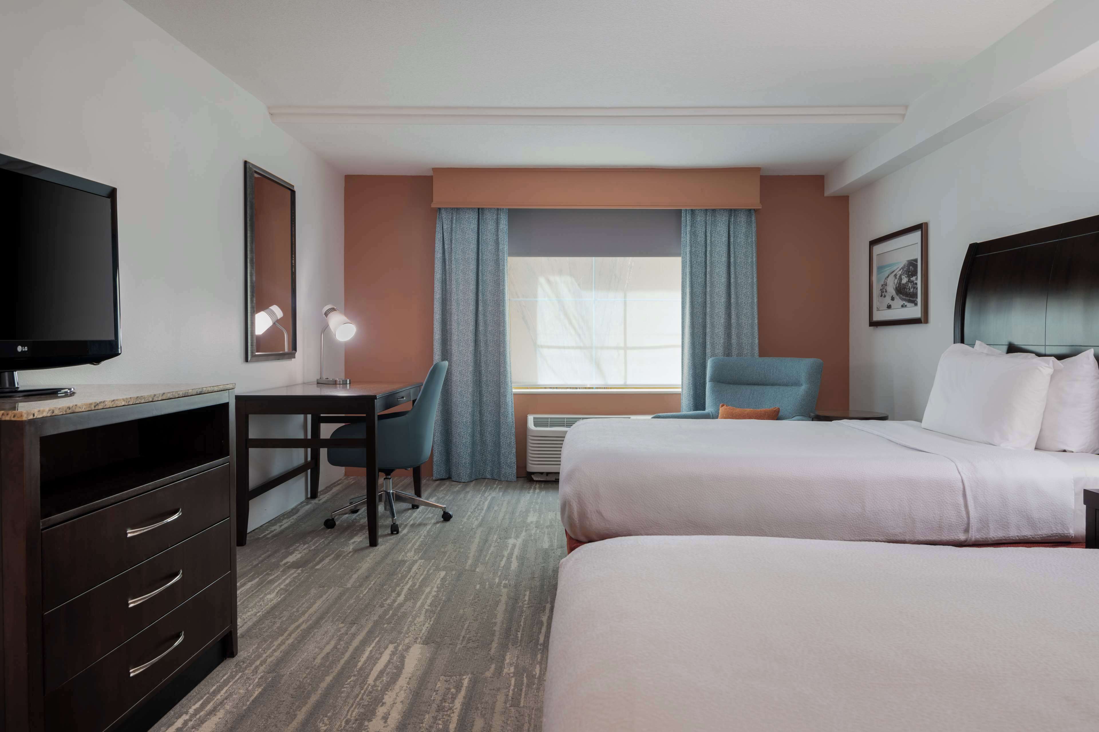 A hotel room with a bed, a desk with a chair, a TV, and a window with curtains.