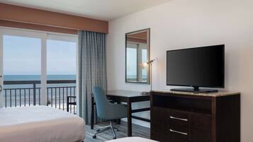 Habitación de hotel con cama, escritorio, silla, televisor y ventana grande con vista al océano.