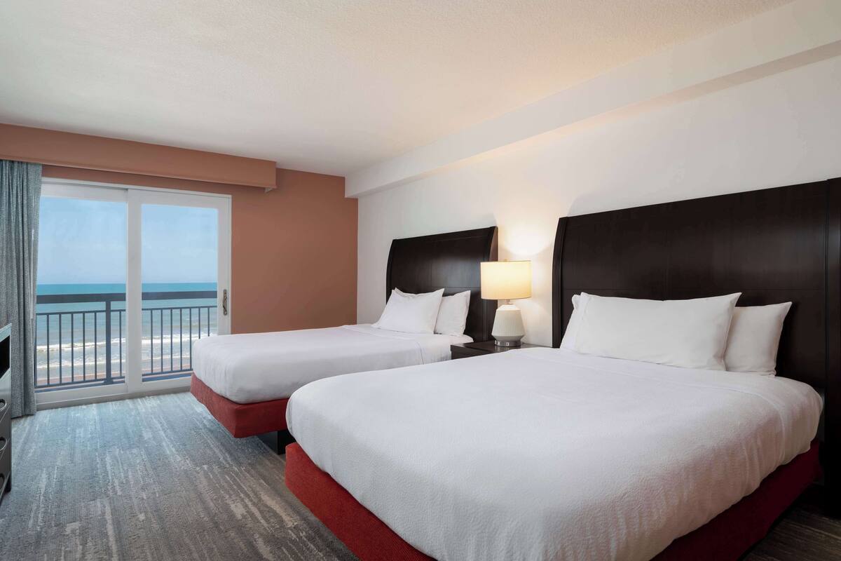 Habitación de hotel con dos camas, un ventanal con vista a la playa y un balcón.