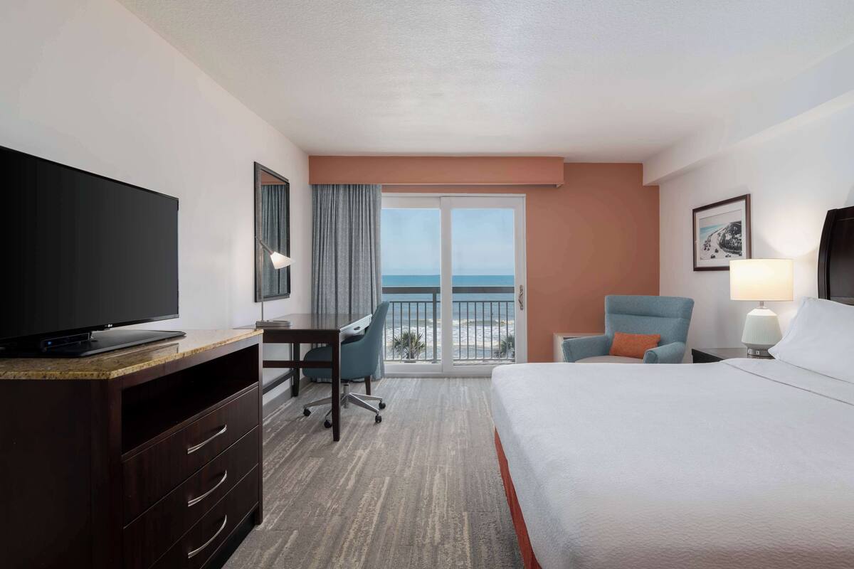 Habitación de hotel con cama, escritorio, silla, televisión, balcón con vista al océano y lámpara.