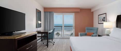 A hotel room with a bed, a desk, a chair, a TV, a balcony with a view of the ocean, and a lamp.