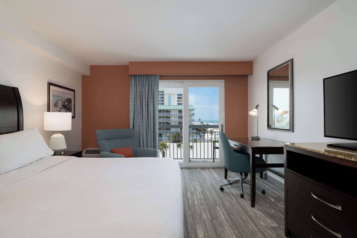 Habitación de hotel con una cama grande, un escritorio, una silla, un televisor y un balcón con vistas a edificios.