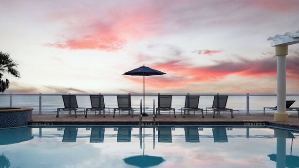 A poolside area with lounge chairs and an umbrella, overlooking the sea with a sunset sky. - Hilton Garden Inn Daytona Beach Oceanfront (Daytona Beach)