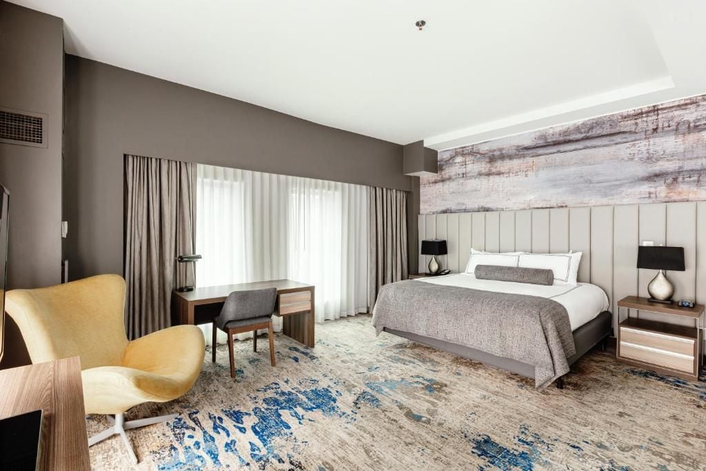 A hotel room with a bed, a desk, a chair, and a large window with curtains.