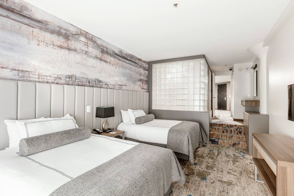 A hotel room with two beds, a large window, a wooden headboard, a bedside table with a lamp, and a TV on a wooden stand.