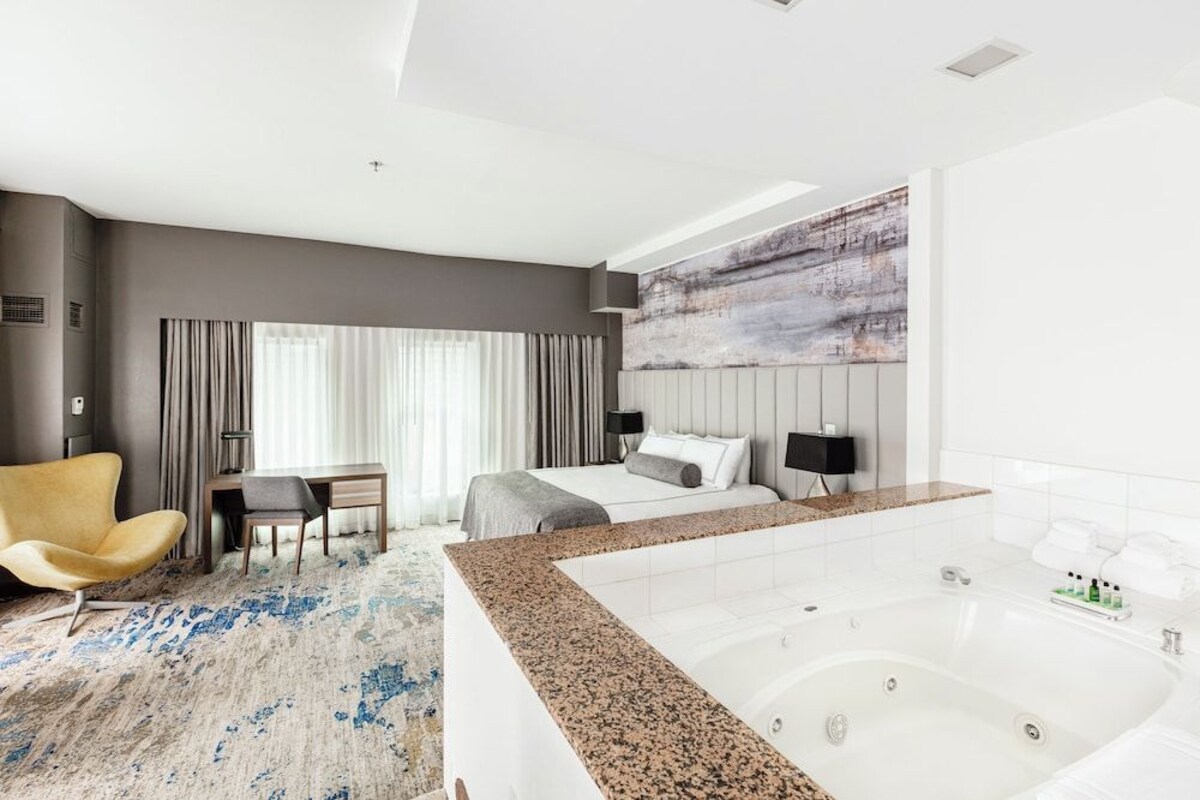 A modern hotel room with a large bed, a sitting area with a chair and table, a large window with curtains, a painting on the wall, and a jacuzzi tub.
