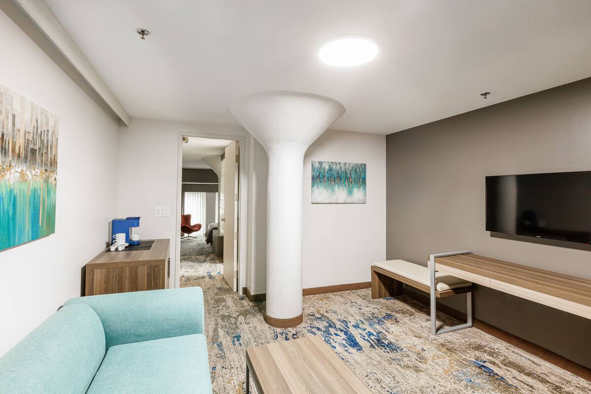 A modern hotel room with a sofa, a coffee station, a TV, and a large pillar.
