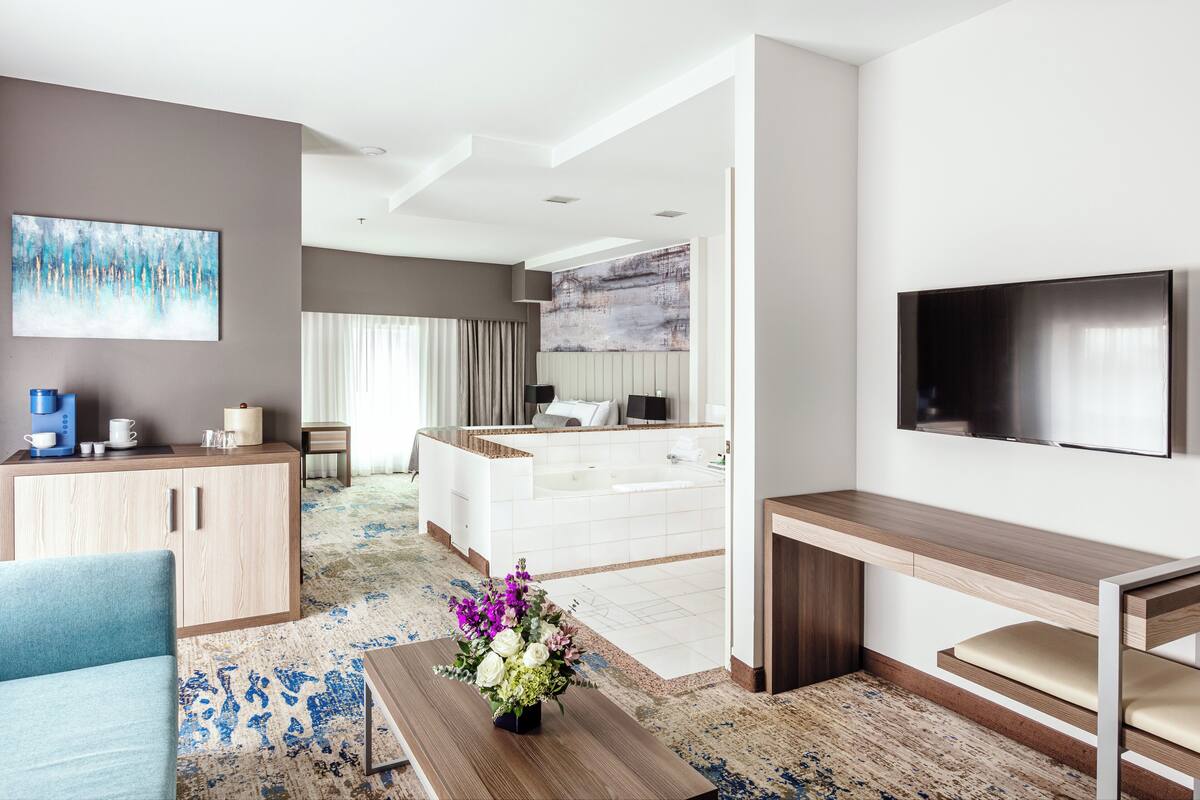A modern hotel room with a large flat-screen TV, a sofa, a coffee station, a bathroom with a bathtub, and a large window with curtains.