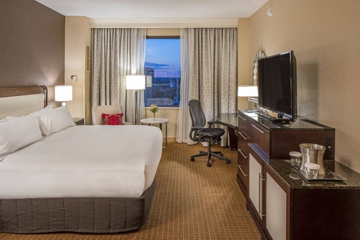 A hotel room with a large bed, a desk with a flat-screen TV, a chair, a small table with a vase, and a view of the city through the window.