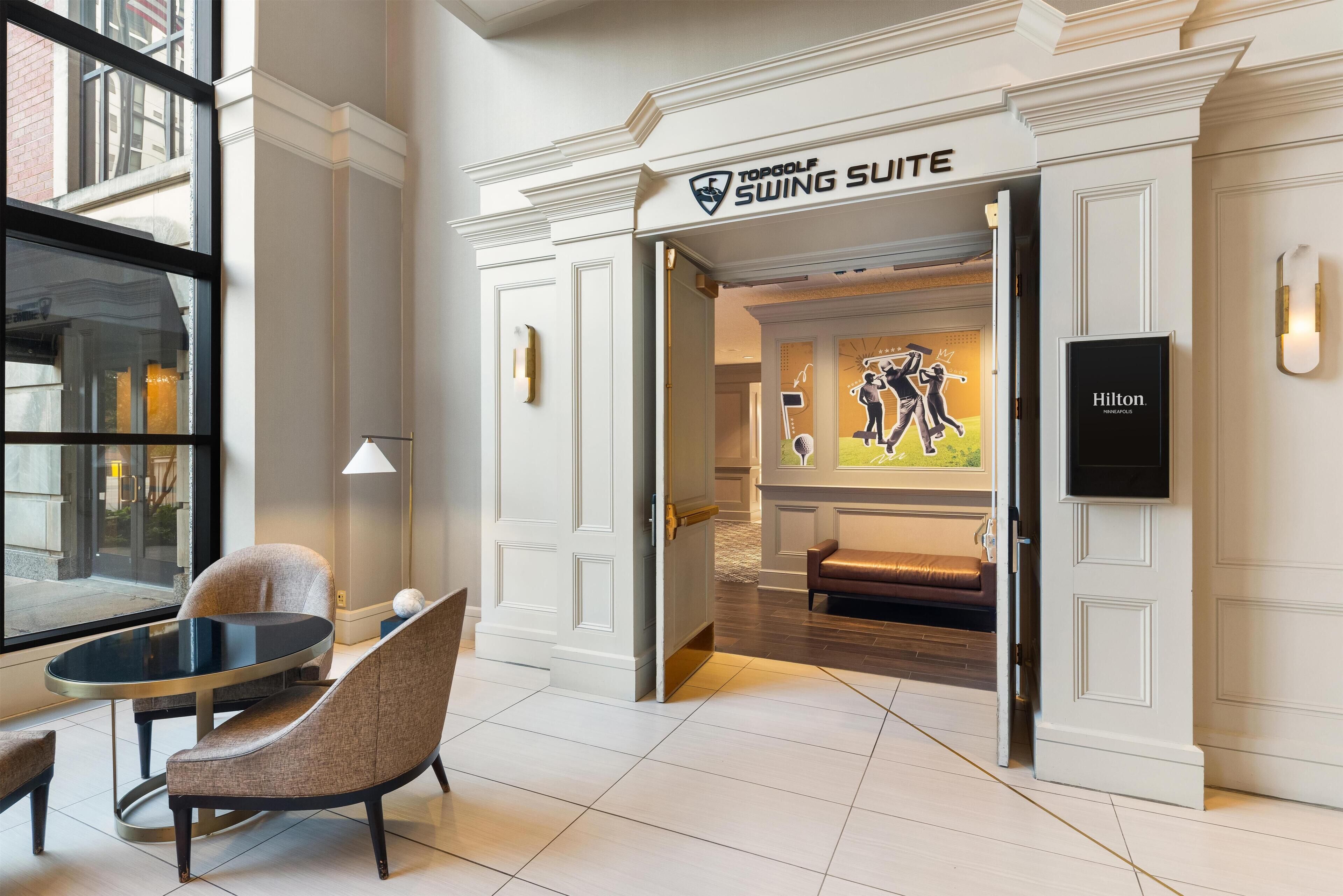 Vestíbulo de hotel con un letrero que dice 'Swing Suite', un área de descanso con mesa y sillas, y vista al interior a través de una puerta abierta.