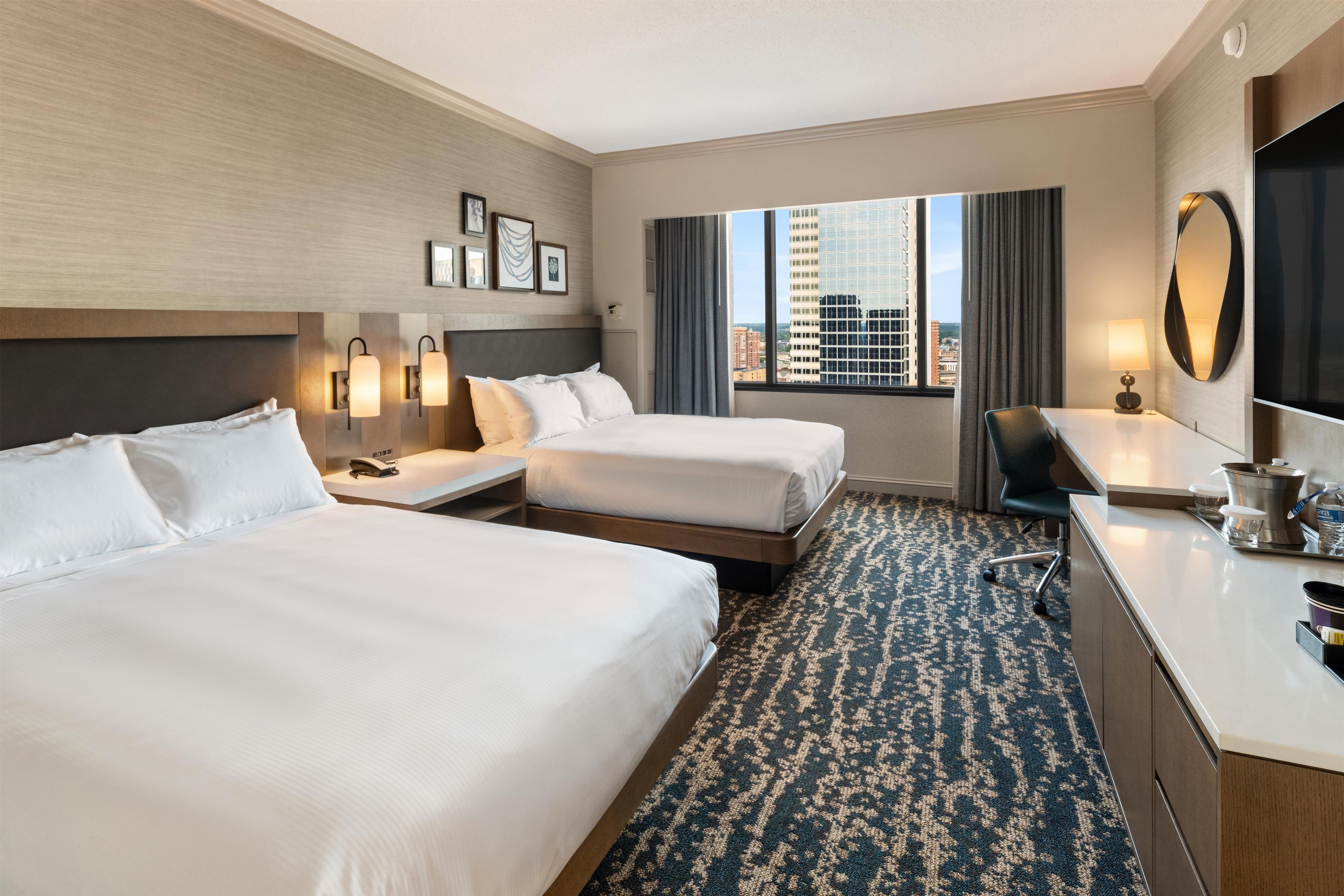A hotel room with two beds, a desk, a chair, a large mirror, and a view of the cityscape through the window.