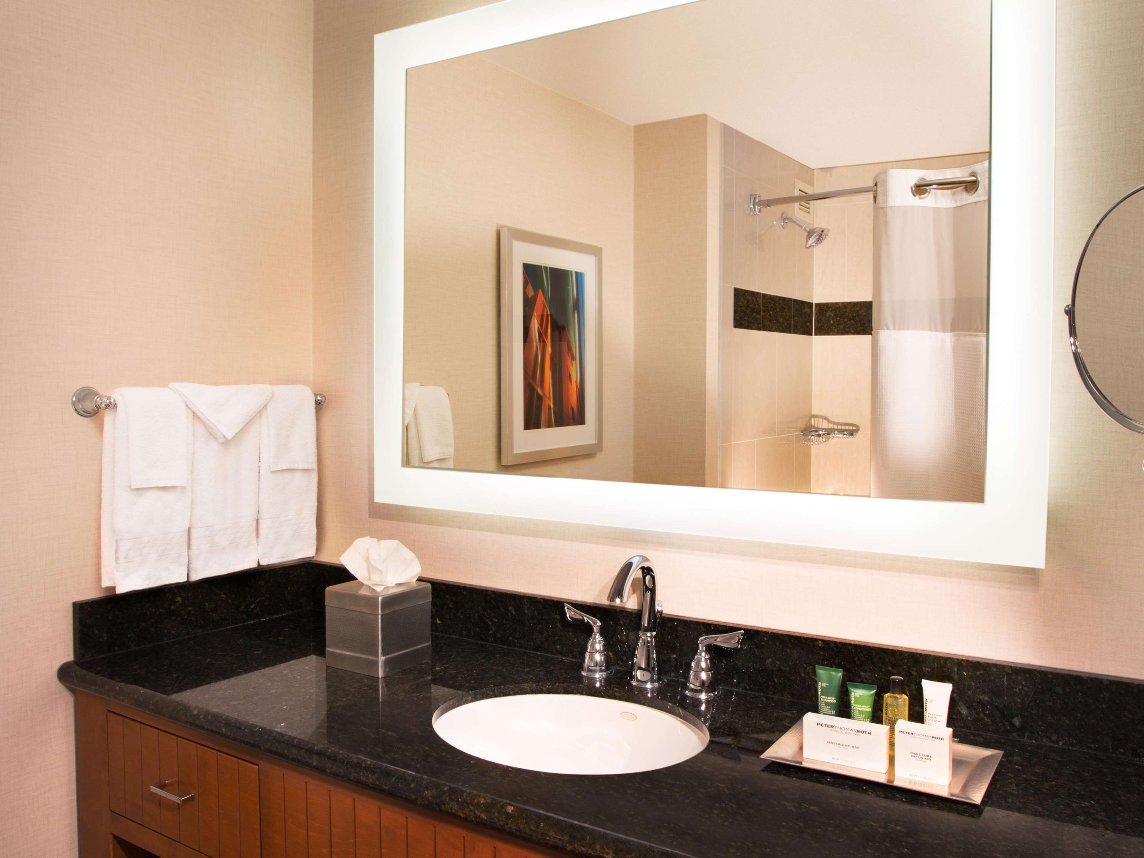 A bathroom with a large mirror, a sink, and a shower. There are towels hanging on a rack and toiletries on the counter.