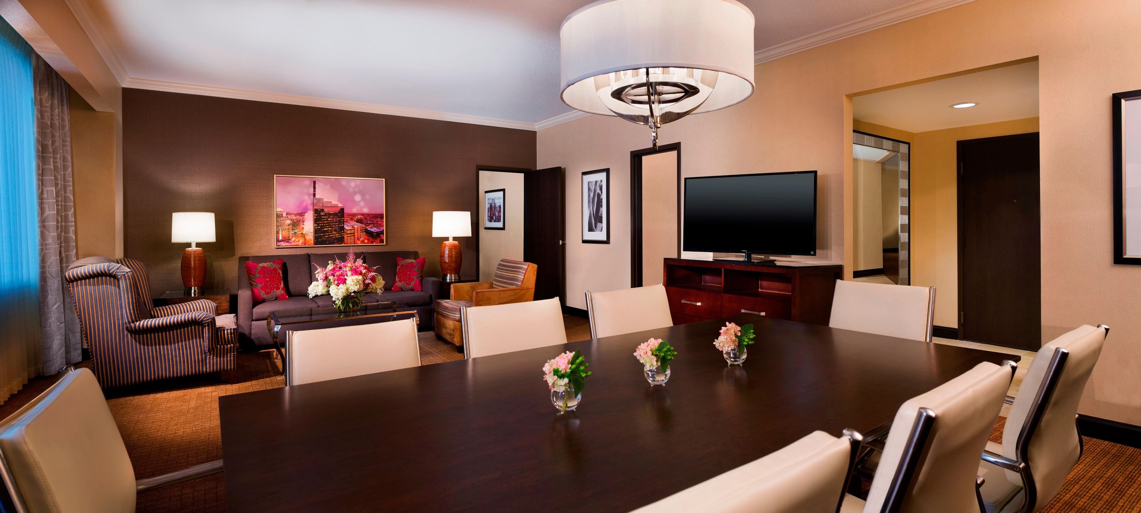 A hotel room with a dark wooden dining table, white chairs, a sofa, a television, and framed artwork on the walls.