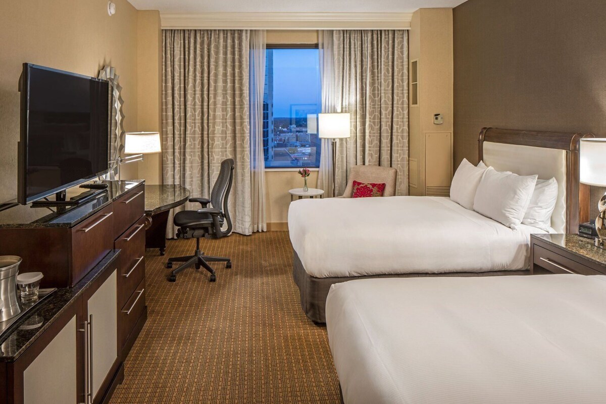 A hotel room with two beds, a desk with a TV, a chair, a window with curtains, and a view of the city.