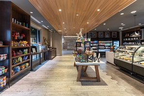 Interior de una tienda con piso de madera, estantes llenos de bocadillos, una mesa de exhibición central y una sección refrigerada a la derecha.