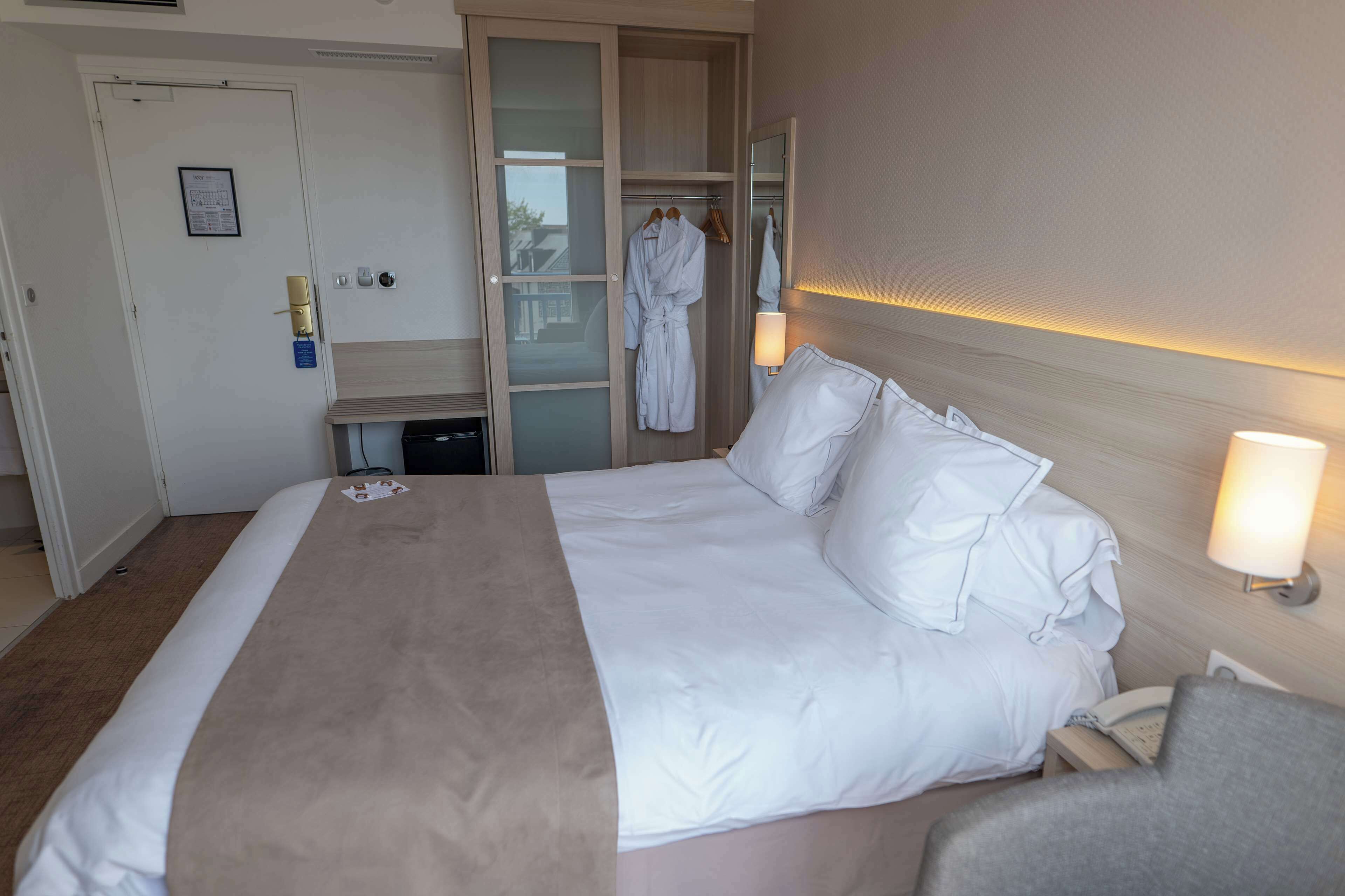 Ein Hotelzimmer mit einem großen Bett, einem Nachttisch, einer Lampe, einem Kleiderschrank und einem Spiegel.