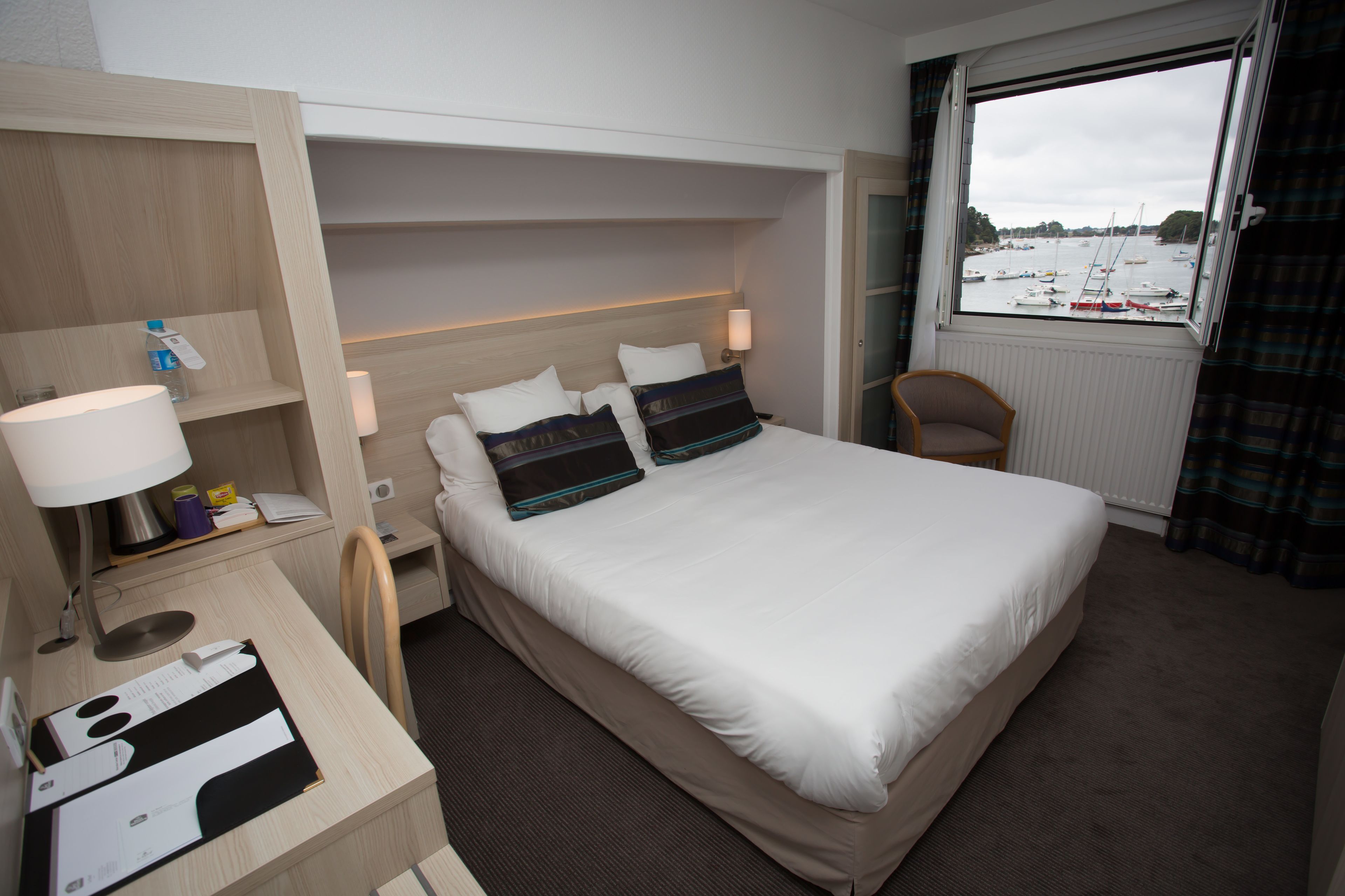 Een hotelkamer met een bed, een bureau met lamp, een stoel en een raam met uitzicht op boten.