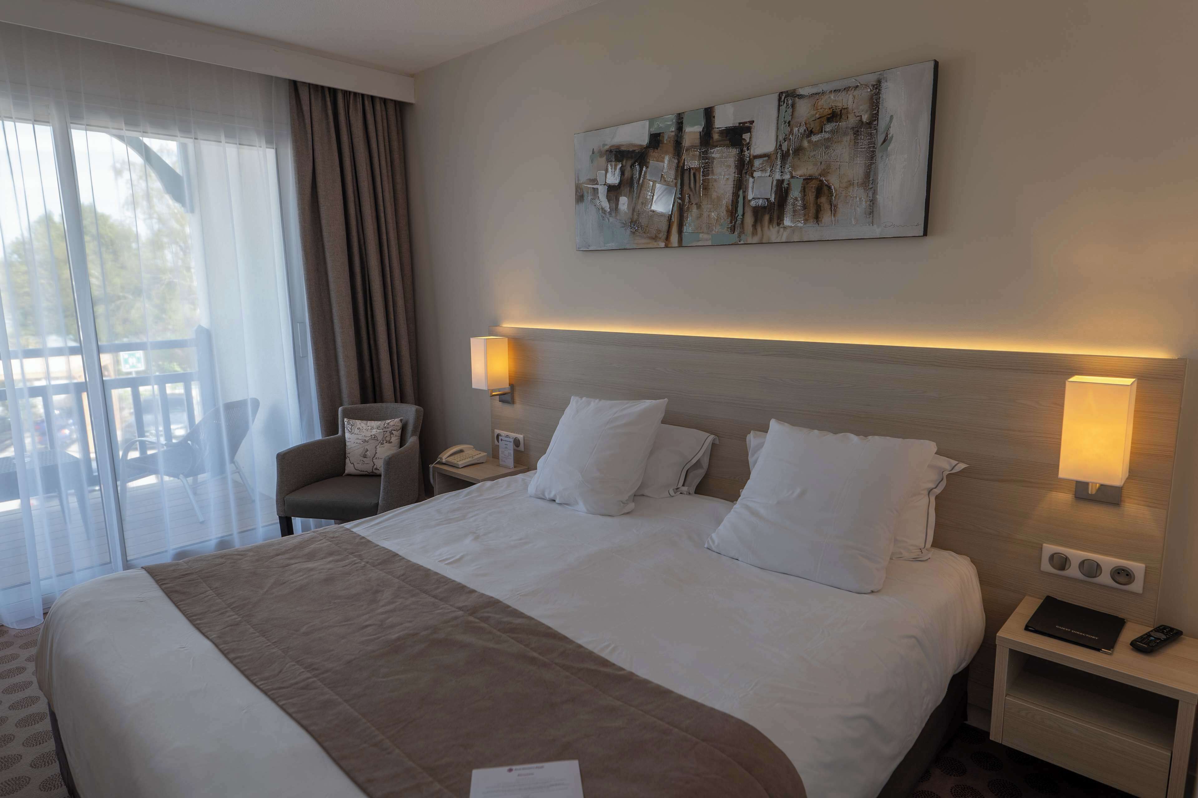 Une chambre d’hôtel avec un grand lit, deux lampes de chevet, une table de chevet avec un téléphone et une télécommande, une chaise et une fenêtre avec des rideaux.