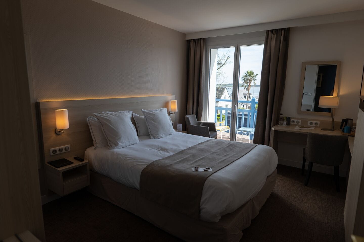 Quarto de hotel com cama grande, abajures, escrivaninha e vista para o exterior.