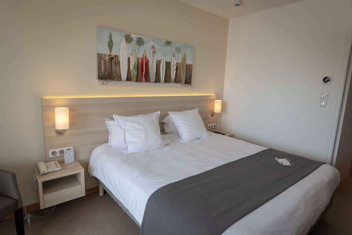 Una habitación de hotel con una cama grande, dos mesitas de noche, una lámpara de noche, un cuadro con tablas de surf y una puerta.