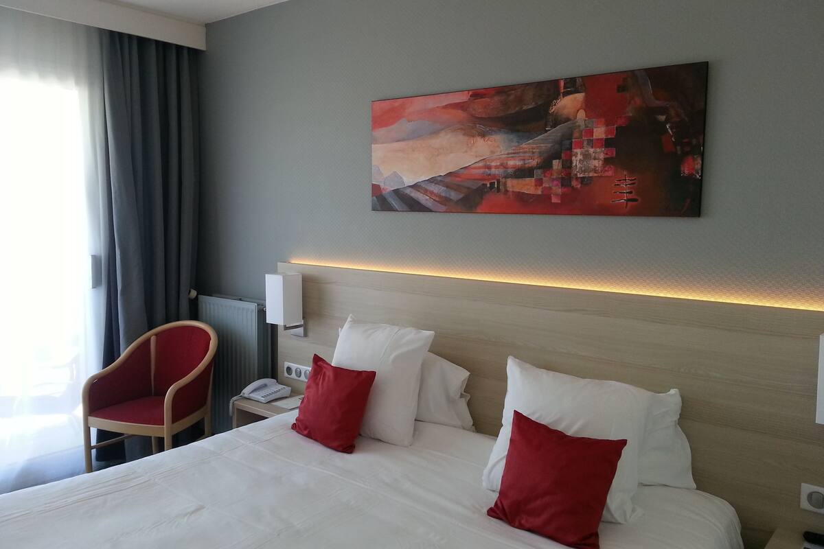 Habitación de hotel con una cama grande, almohadas rojas y blancas, una silla roja, un cuadro en la pared y una mesita de noche con un teléfono.