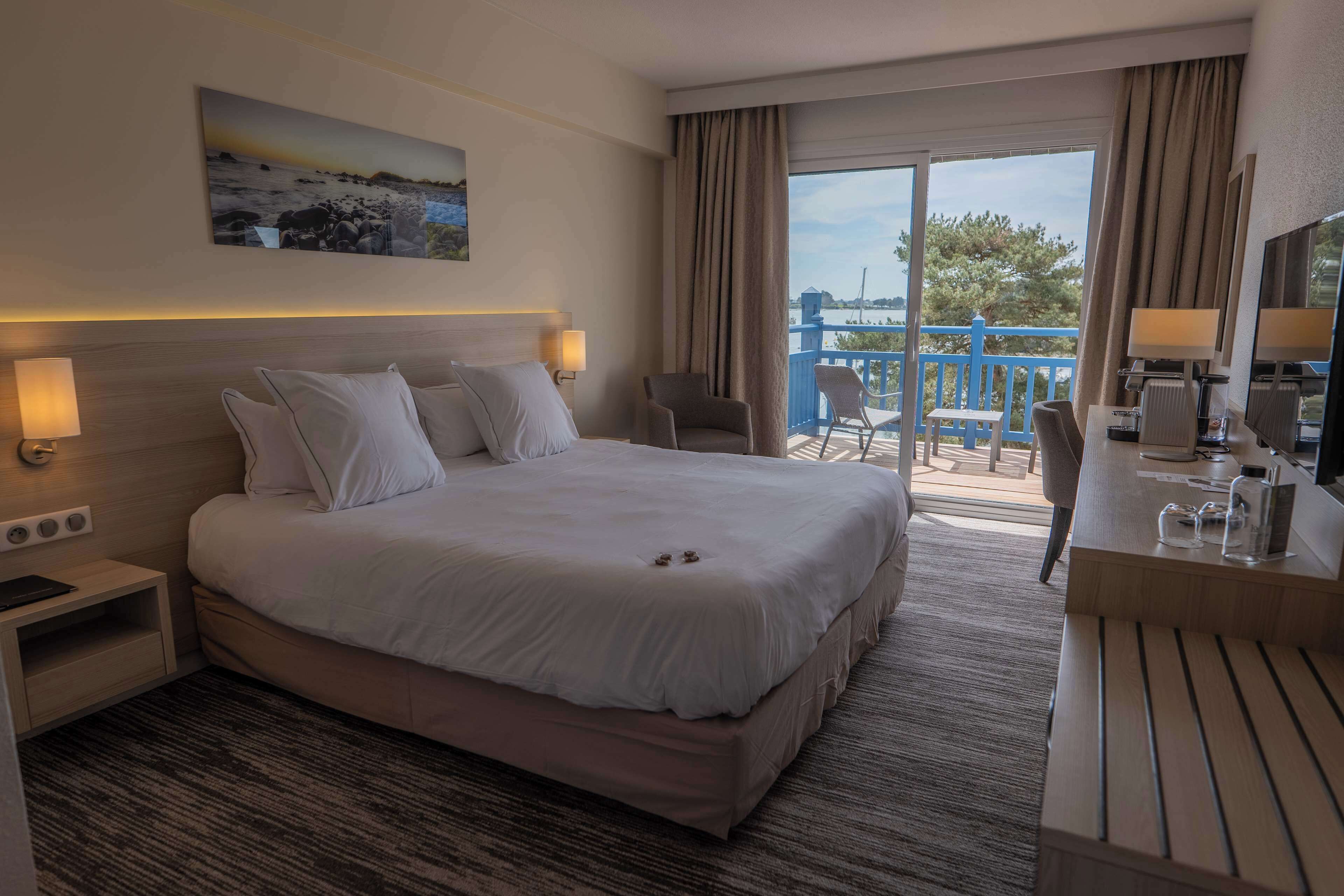Habitación de hotel con una cama grande, un escritorio con silla, una mesita de noche con lámpara y vistas al balcón exterior.