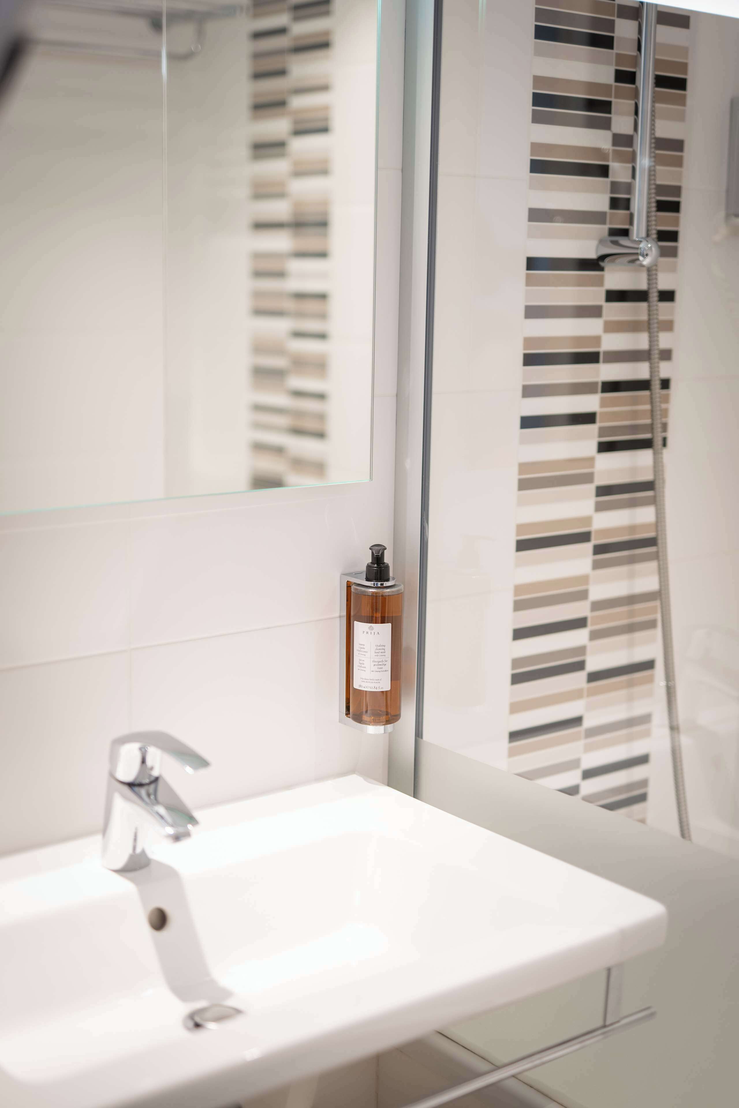 A modern bathroom with a glass shower enclosure, a white sink, and a bottle of hand soap on the wall.
