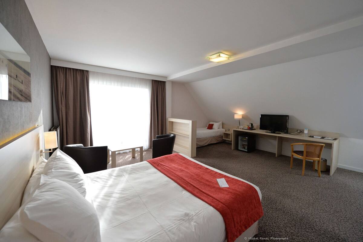 Habitación de hotel con cama, escritorio, silla, televisor y ventana con cortinas.