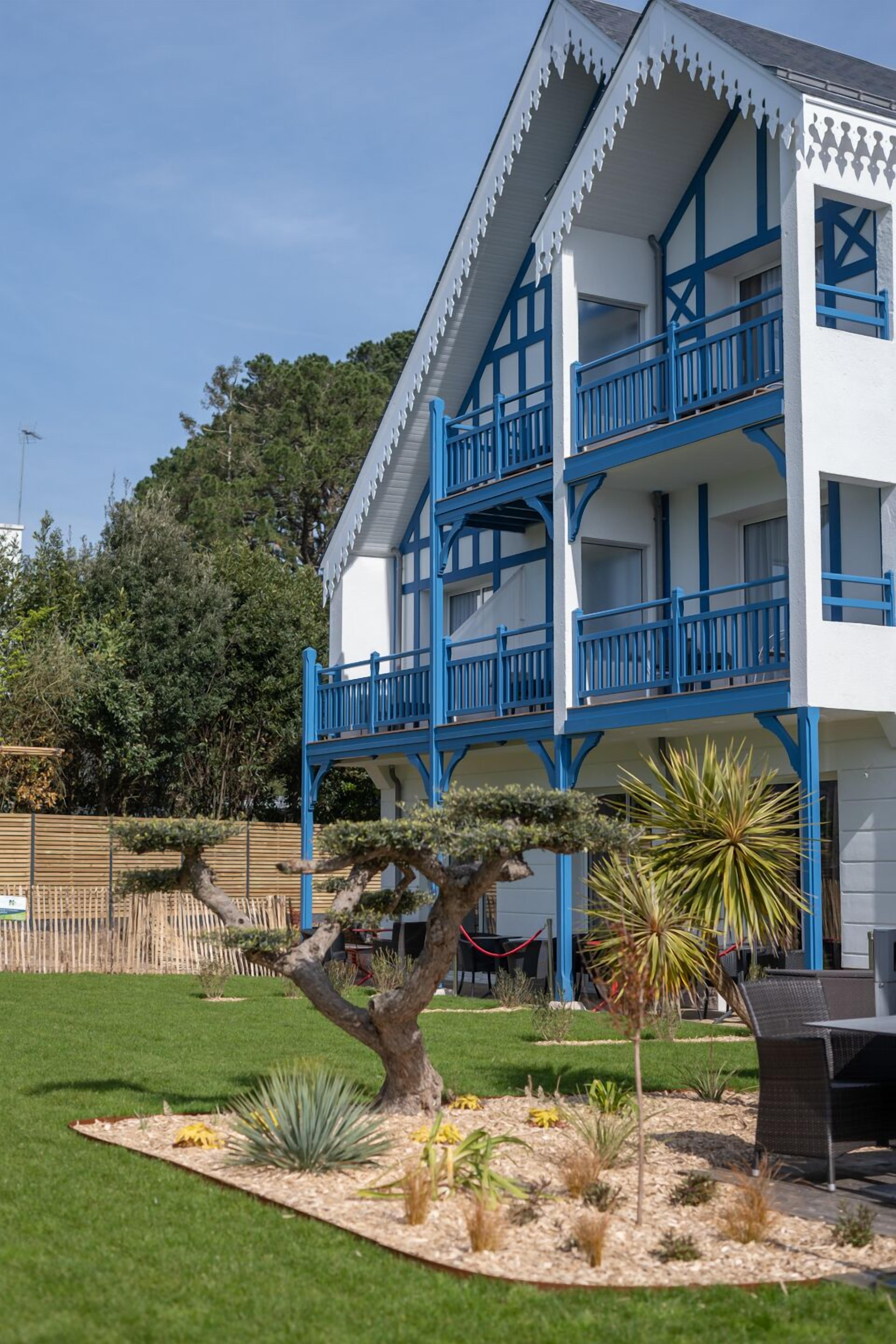 A two-story building with a white facade and blue accents, featuring balconies and a well-maintained garden with various plants.