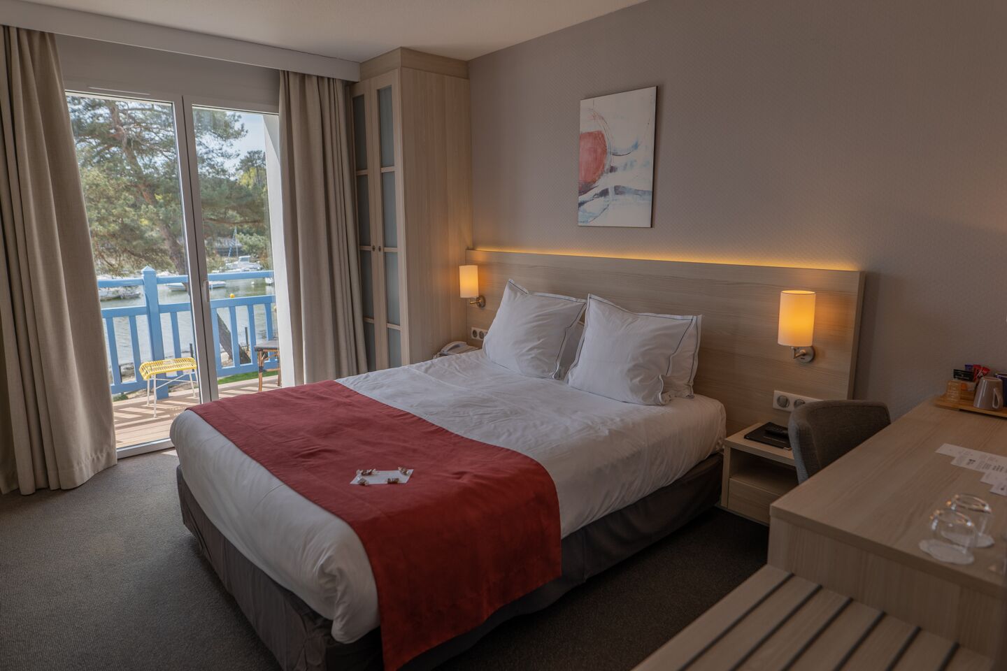 Habitación de hotel con cama, mesitas de noche, escritorio y vista a un espacio exterior azul.