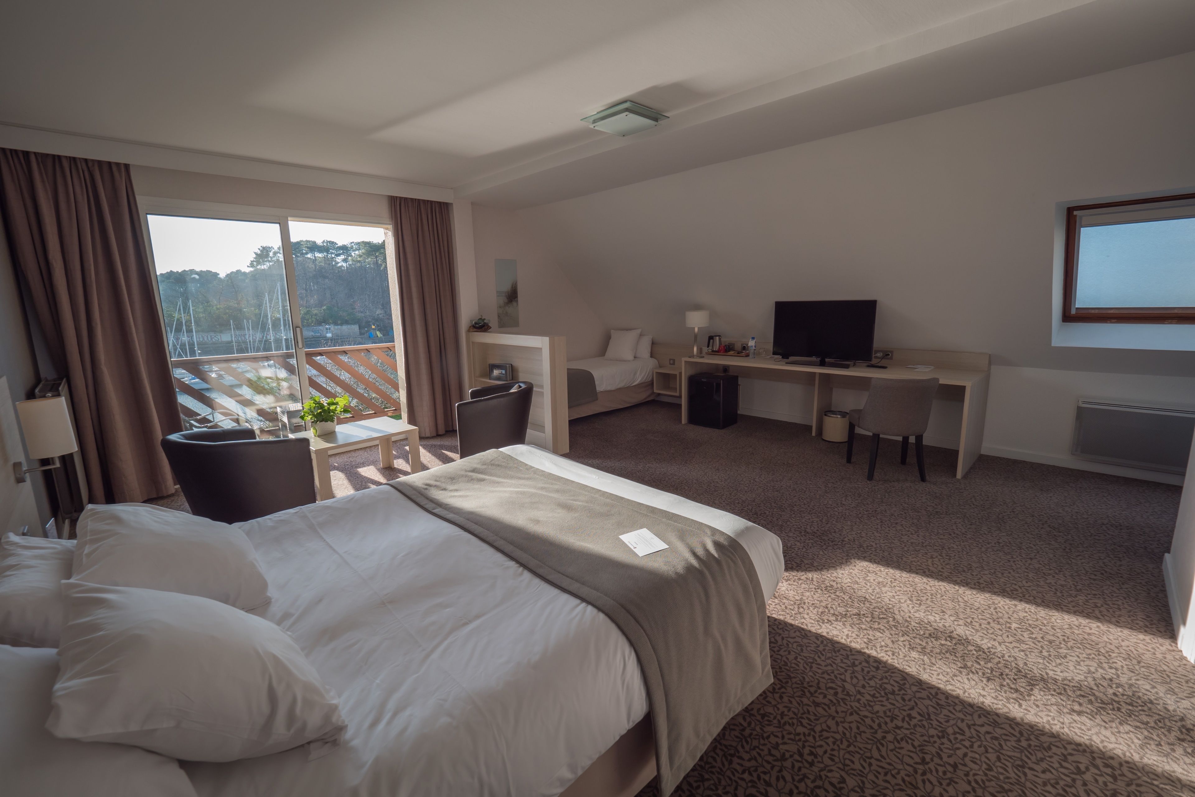 Ett hotellrum med en säng, ett skrivbord, en TV och en balkong med utsikt över träd.