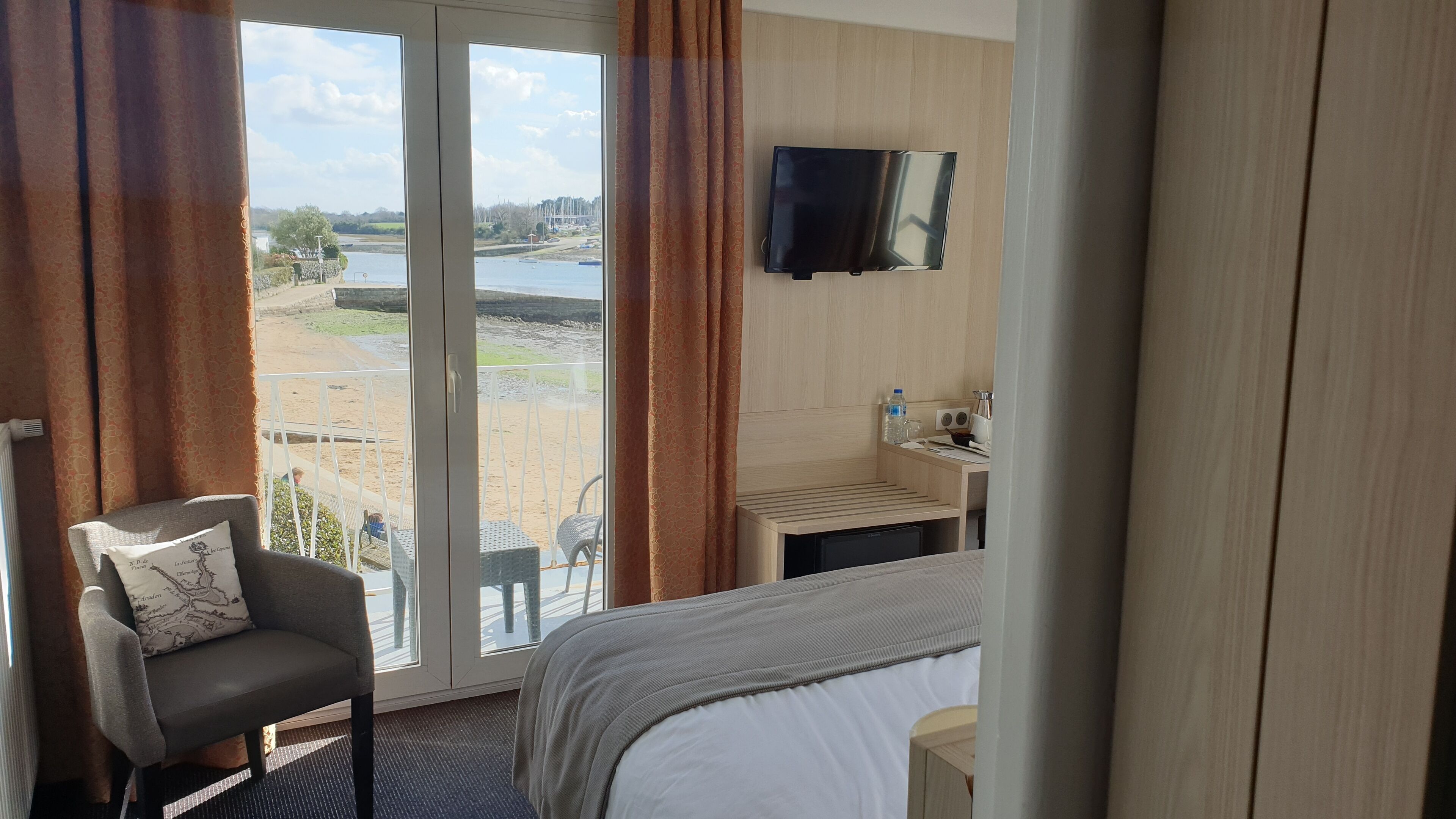 A hotel room with a bed, a chair, a TV, a small table with a sink, and a view of a beach and water.