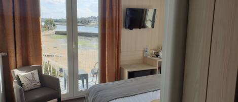 A hotel room with a bed, a chair, a TV, a small table with a sink, and a view of a beach and water.