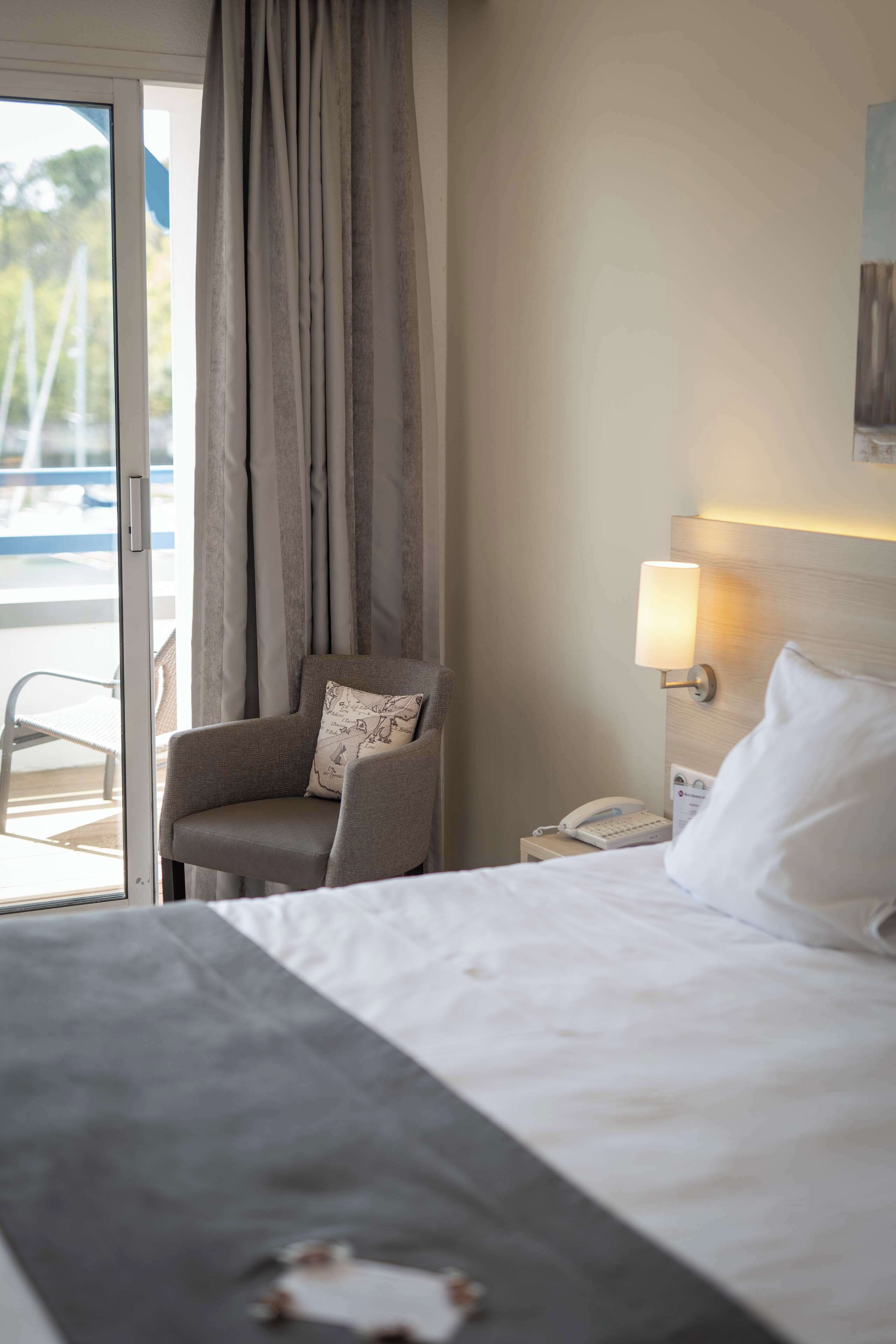 A hotel room with a bed, a chair, a nightstand with a lamp, and a view of the outdoors through a sliding door.