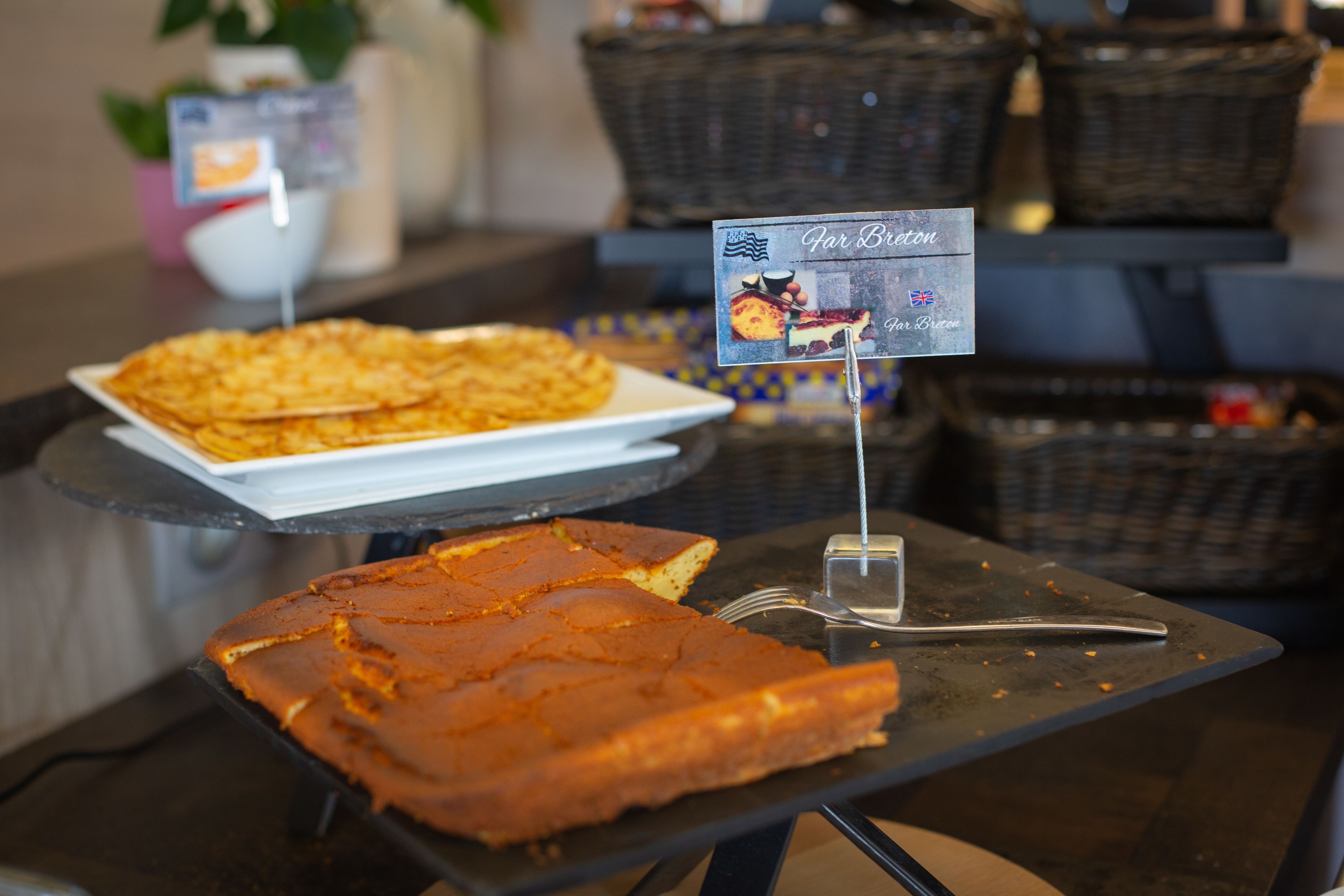 A buffet table with a variety of food items, including a large piece of cake and a plate of what appears to be a casserole or casserole-like dish. There are also baskets in the background, possibly containing additional food items.